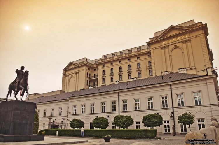 Hotel Bristol wraz z pomnikiem księcia Józefa Poniatowskiego. A nad Nimi delikatnie przebijające się przez chmury, poranne słońce.
